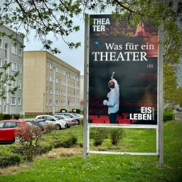 In einer Plattenbausiedlung steht ein Werbeplakat des Theater Eisleben, auf dem eine Person in einem Theaterraum steht und eine einzelne Rose hochhält.