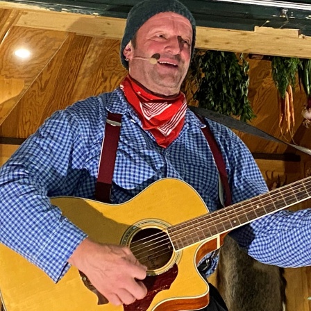 Der Komiker Fidelius Waldvogel aus dem Schwarzwald steht mit der Gitarre auf einer Bühne und singt in ein Mikrofon rein.