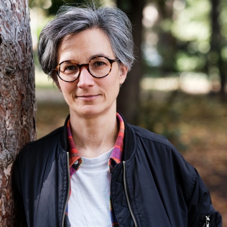 Eine Frau mit dunkelgrauen kurzen Haaren und Brille lehnt an einem Baumstamm und schaut in die Kamera.