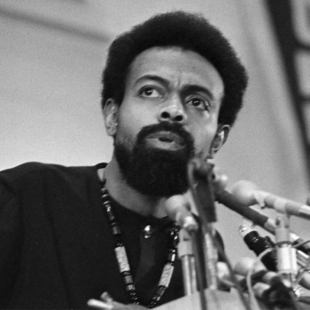 Amiri Baraka spricht an einem Pult in mehrere Mikrofone