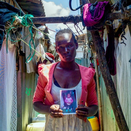Die Mutter Lumbasi hält ein Foto ihres dreijährigen Sohnes in die Kamera und steht vor einer Hütte in einem Slum in Kenia.