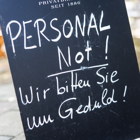 Auf einer Tafel am Eingang zu einem Biergarten ist der Schriftzug "Personalnot! Wir bitten Sie um Geduld!" zu lesen.