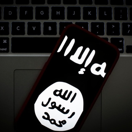 Das Logo des islamischen Staates auf dem Bildschirm eines Smartphones. Im Hintergrund ein Laptop.