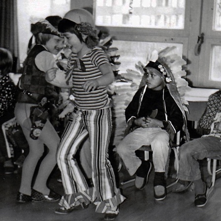 Verkleidete weiße Kinder feiern Fasching in einem Klassenzimmer. Zwei Kinder tanzen, ein schwarzer Junge im Indianerkostüm sitzt auf einem Stuhl daneben.