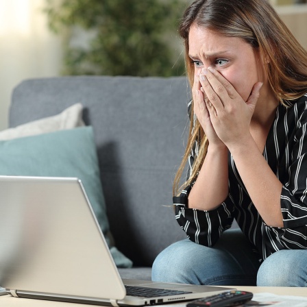 Eine Frau sitzt betroffen vor einem Laptop und hält sich die Hände vor Mund und Nase