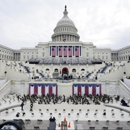 Blick auf das Kapitol in Washington mit herabhängenden großen US-Fahnen und einer probenden Militärkapelle, davor mit großem Abstand leere Stühle