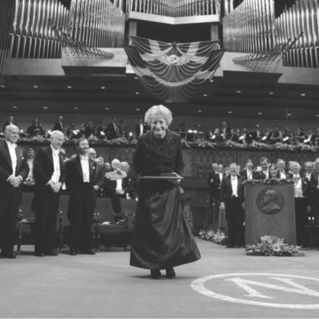 Das Schwarz-Weiß-Foto zeigt eine sanft lächelnde ältere Frau, eine Urkunde haltend beim Durchschreiten des Konzerthauses Stockholm