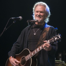 Sänger und Songwriter Kris Kristofferson steht lächelnd auf einer Bühne mit einer Akustikgitarre in der Hand und einer Mundharmonika vor sich.