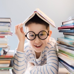 Ein Junge sitzt zwischen 2 Bücherstapekn mit einem Buch auf dem Kopf