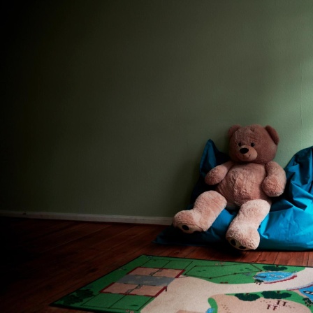 Ein großer Teddy sitzt auf einem Sitzsack in einem Spielzimmer. Daneben liegen weitere Spielsachen.