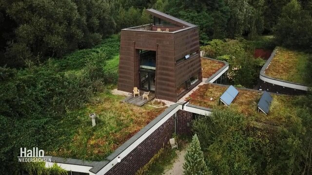 Ein turmähnliches Haus aus dunklem Holz auf einem Backsteinhaus mit Grasdach, umgeben von einem Park.