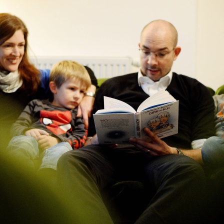 Mann und Frau sitzen mit Kindern gemütlich auf ihrem Sofa und lesen gemeinsam ein Buch
