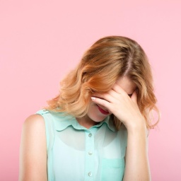 Eine Frau in mintgrüner Bluse vor pinkem Hintergrund, hält sich vor Scham die Hand vor das Gesicht.