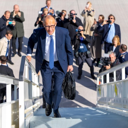 Friedrich Merz läuft die Gangway eines Fliegers hinauf. Unter ihm am Boden stehen Fotografen und fotografieren zu ihm hinauf.