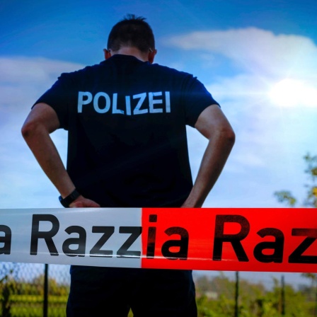 Ein Polizist steht vor blauem Himmel mit dem Rücken zur Kamera. Davor Absperrband mit der Aufschrift Razzia.