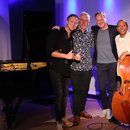 Vier Männer lächeln freundlich, sie stehen zwischen mehreren Musikinstrumenten.