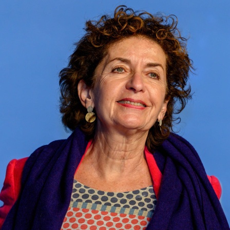 Ruth Beckermann steht vor einem blauen Hintergrund, trägt einen roten Mantel und einen blauen Schal über einem farbenfrohen Oberteil.