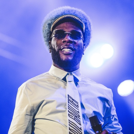 Burna Boy, mit einem Weißen Hemd, Fellkappe, Sonnenbrille  und Schlips auf der Bühne. 