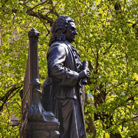 Johann Sebastian Bach: Denkmal vor der Leipziger Thomaskirche im Profil. Im Hintergrund satt begrünte Bäume.