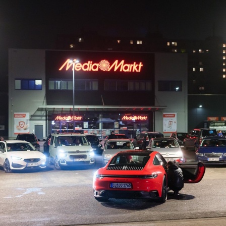 Autolesung auf einem Media-Markt-Parkplatz im Rahmen der Grand Snail Tour. Ein roter Porsche 911 Carrera steht zentral vor einer Ansammlung weiterer PKWs.