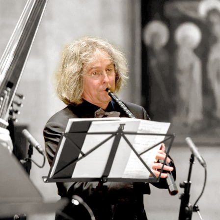 Arno Paduch (in Farbe) steht mit Instrument vor Notenständer, Vordergrund und Hintergrund (schwarzweiß) zeigen Fragmente von Ensemble