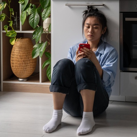 Eine traurige Frau sitzt auf dem Boden und schaut aufs Smartphone. (Bild: picture alliance / Zoonar | Dmitrii Marchenko)