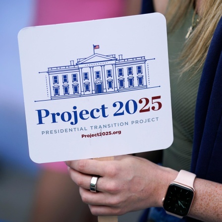Eine Person hält ein Schild mit der Aufschrift "Project 2025" in der Hand. Darauf ist außerdem eine Zeichnung des Weißen Hauses zu sehen.