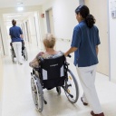 Zwei Pflegerinnen schieben Patient:innen im Rollstuhl auf einem Flur in einem Krankenhaus