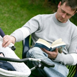 Vater sitzt mit einem Buch im Garten und betreut seinen sieben Wochen alten Sohn, der im Kinderwagen liegt.