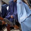 Ein Taliban-Kämpfer in Kabul hält eine Kalaschnikow, während im Hintergrund Frauen in Burkas sitzen.