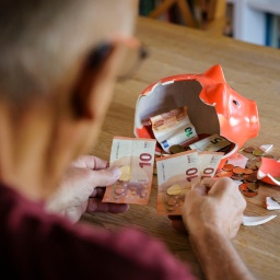 Ein alter Mann sitzt zu Hause am Tisch vor einem zerbrochenen Sparschwein und zählt Geld.
