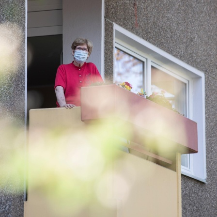 Eine Seniorin mit Coronamaske steht auf ihrem Balkon.