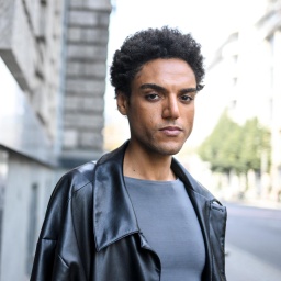 Lamin Leroy Gibba, er trägt eine schwarze Jacke und ein graues T-Shirt. Der Hintergrund ist verschwommen.