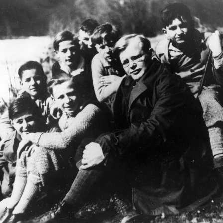 Dietrich Bonhoeffer sitzt inmitten einer Gruppe Jungen an einem Flussufer, alle schauen in die Kamera.