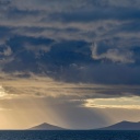 Stürmische dunkle und helle Wolken über flachen Inselkuppen im Meer mit einem Hauch von Sonne.