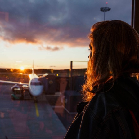 Junge Frau blickt bei Sonnenuntergang aus Flughafenfenster auf Flugzeug auf dem Rollfeld