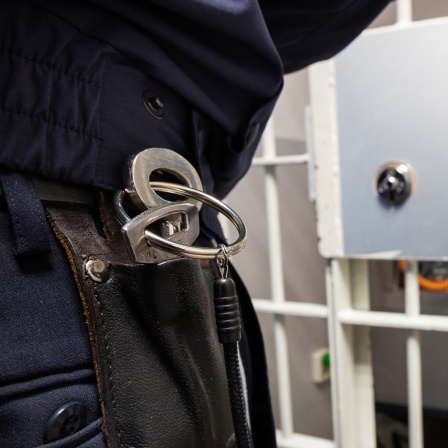 Blick durch die Gittertür in einer JVA. Vor der Gittertür ist eine Person zu erahnen mit Schlüsselbund, dahinter ein Gefängnisflur.