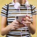 Eine Frau steht mit Smartphone in der Hand und Kopfhörern vor einer gelben Wand