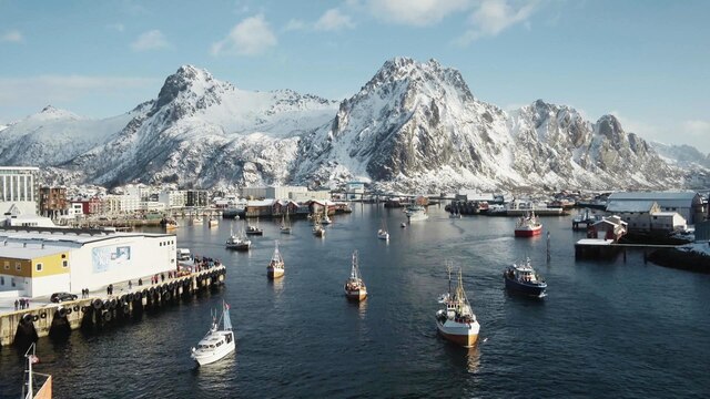 Mehrere Fischerboote fahren aus einem kleinen Hafen, im Hintergrund sind verschneite Berge zu sehen.