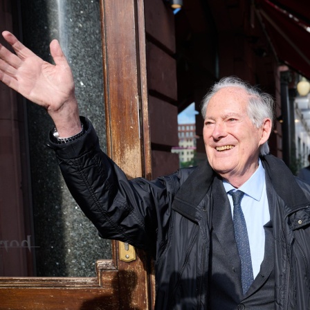 Der Unternehmensberater Roland Berger steht vor einem Restauranteingang, lacht und winkt in die Kamera. Er trägt eine schwarze Windjacke über seinem Anzug.