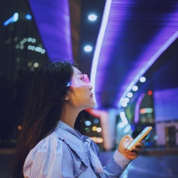 Eine junge Frau steht unter einer Brücke und nutz allemöglichen technischen Geräte. Eine Datenbrille und eine Art Smartphone. Im Hintergrund sind die Lichter der Stadt bei Nacht zu erkennen. Die Brücke unter der sie steht ist blau erleuchtet.