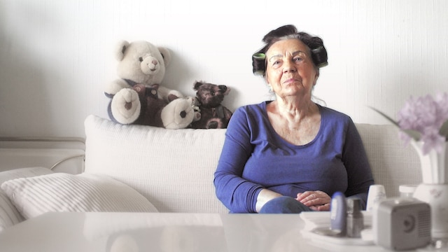 Rentnerin Christel C. sitzt mit Lockenwicklern auf ihrem Sofa. Auf der Sofalehne sitzen zwei Teddybären.