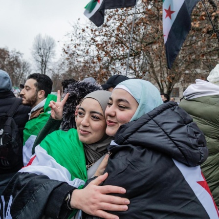 Zwei junge Frauen, teilweise in eine syrische Flagge gehüllt, umarmen sich in einer Menschenmenge in Berlin.