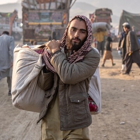Ein geflüchteter Afghane steht in Pakistan an einem Grenzübergang. Er trägt einen Gegenstand, der in ein Tuch eingehüllt ist und blickt dabei in die Kamera.