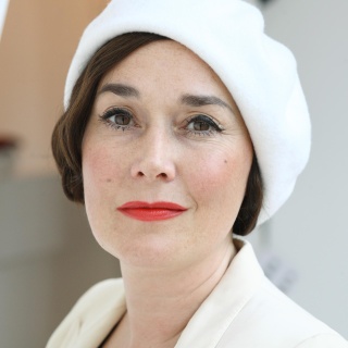 Porträtfoto einer weißen Frau mit kurzen, braunen Haaren, braunen Augen und rot geschminkten Lippen. Sie trägt eine elegante weiße Mütze, die leicht schräg auf ihrem Kopf sitzt. Es handelt sich um Fiona Bennett.