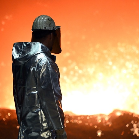 Ein Mann mit Schutzkleidung steht an einem Hochofen und sieht ins Feuer
