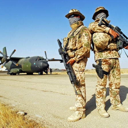 Zwei Soldaten der Bundeswehr stehen vermummt in einer Wüstenlandschaft. Im Hintergrund ein Transportflugzeug.