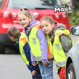 Drei Kinder mit Warnwesten schauen am Straßenrand nach Autos