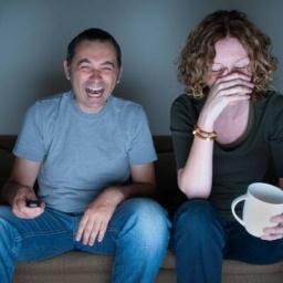Ein Mann und eine Frau sitzen auf der Couch vor dem Fernseher und lachen.