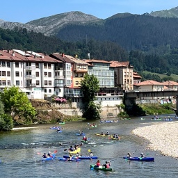 Menschen paddeln in Booten auf einem Fluss in Asturien, im Hintergrund eine Stadt, eine Brück und Berge 